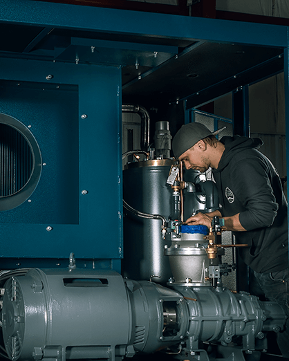 man-wearing-a-hat-backwards-while-working-on-a-grey-K-Series-oil-lubricated-air-compressor-at-Rogers-Machinery's-Centralia-Washington-production-facility