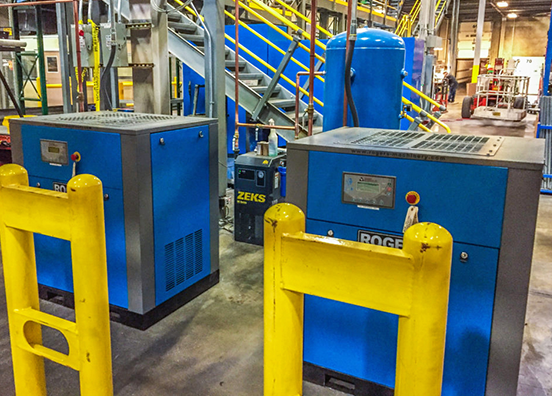 Two-blue-Rogers-Machinery-Company-oil-lubricated-rotary-screw-air-compressor-installed-at-a-customer-sitecustomers-site-in-front-of-an-air-compressor-system-with-a-tank-and-dryer