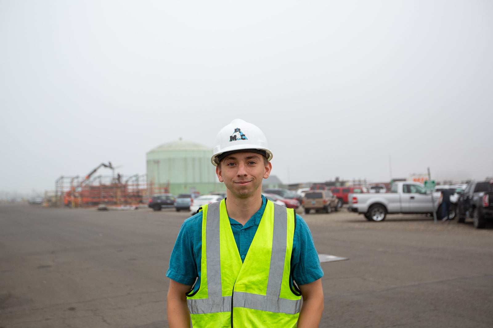 a man in a hard hat and safety vest smiles at the camera in an industrial park